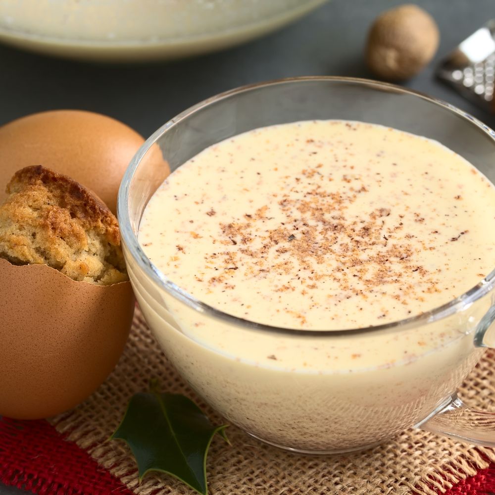 Image 2 article de blog eggnog Le Club des Connaisseurs. Verre de lait de poule crémeux saupoudré de noix de muscade, accompagné d’un biscuit dans une coquille d’œuf, sur fond de nappe festive boisson traditionnelle de Noël.